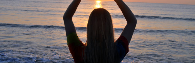 Woman making a heart shape with her hands in front of the ocean with a sunset in the background