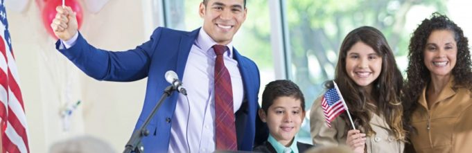 Mid adult Hispanic man is standing on stage with his wife and two children. Candidate is introducing his family to audience while running for local government office.