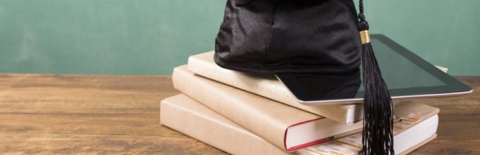 Spring graduation time. A black cap/motarboard with black tassel lies on top of a stack of textbooks and digital tablet. All objects lie on a rustic wooden desk with green chalkboard in background. Copyspace to side. No people. Education themes.