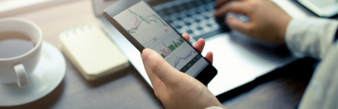 Man checking stocks on his phone while he types on a laptop