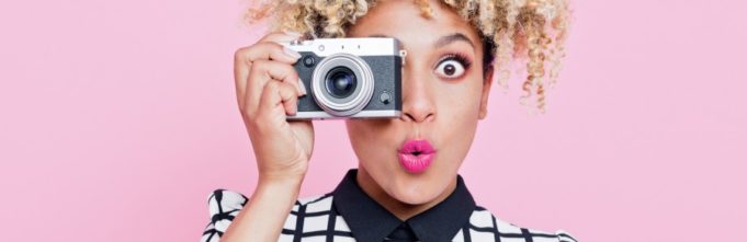 Portrait of surprised beautiful afro american young woman wearing grid check playsuit, holding a camera in hand, looking at camera. Studio shot, one person, pink background.