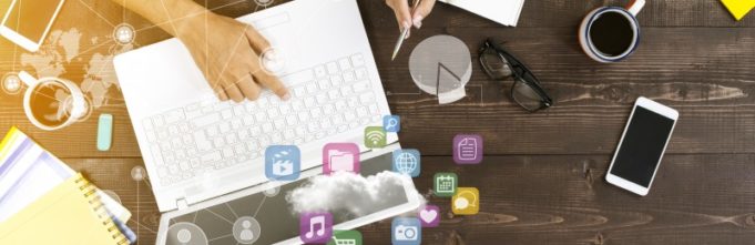 White computer sitting on wooden desk alongside two peoples hands, coffee mug, iphone, glasses, and icons floating above the computer